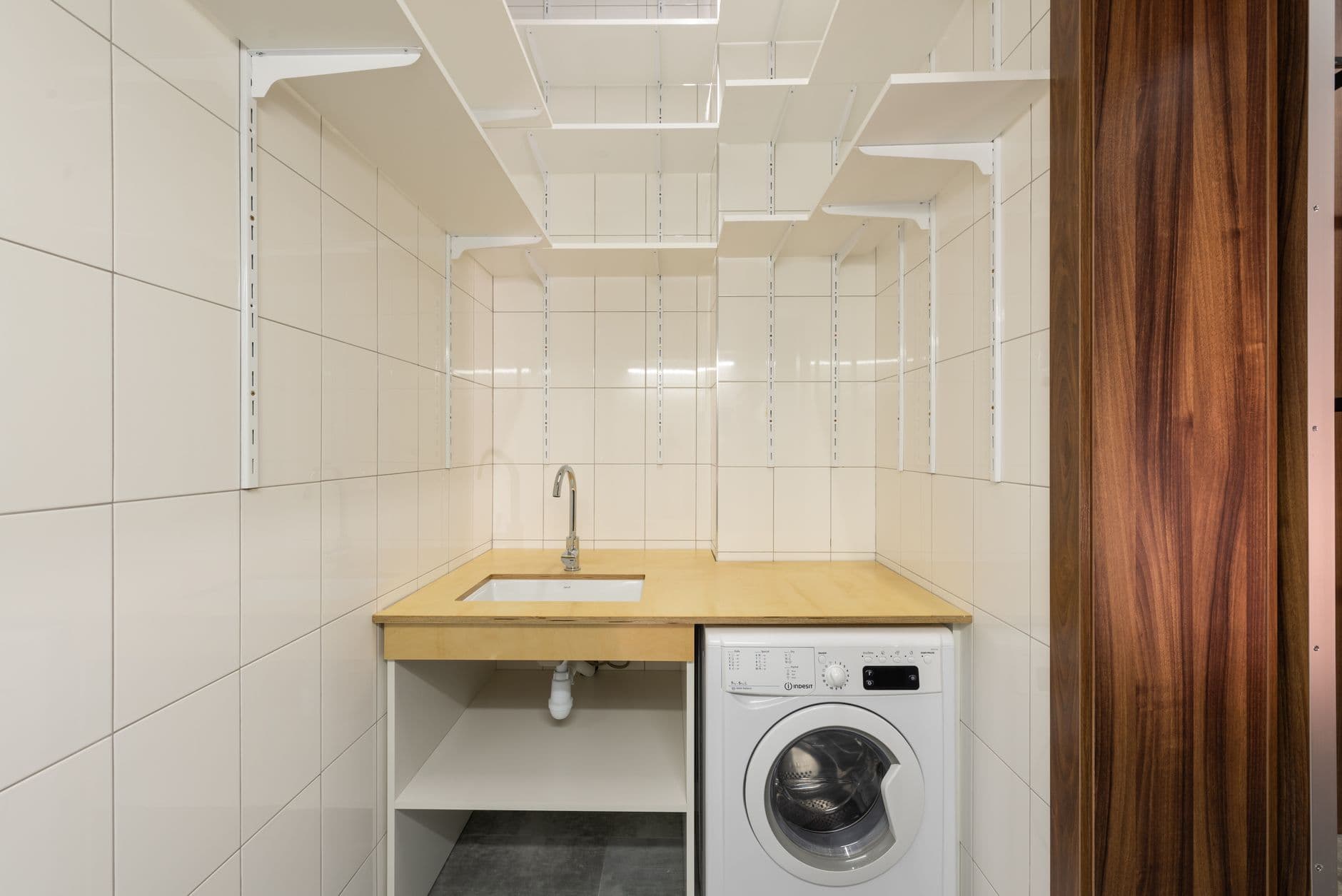 Narrow tiled laundry with a washing machine under a benchtop and plumbing visible below the sink